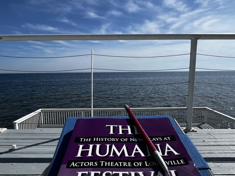 A copy of The Humana Festival resting on the deck railing, with Lake Superior stretching out to the horizon under a blue sky