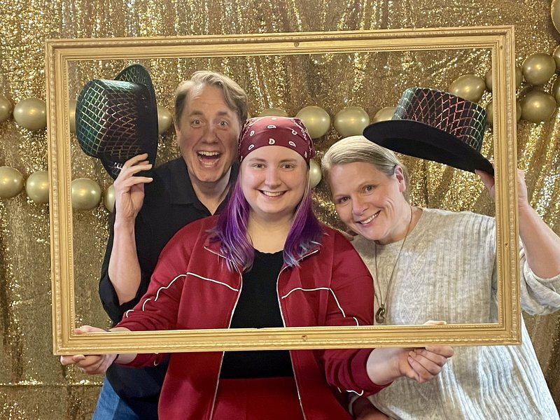 Justin, Kaylee, and Jody posing inside a gold picture frame in front of a gold sequin backdrop, with Justin and Jody holding up sparkly top hats