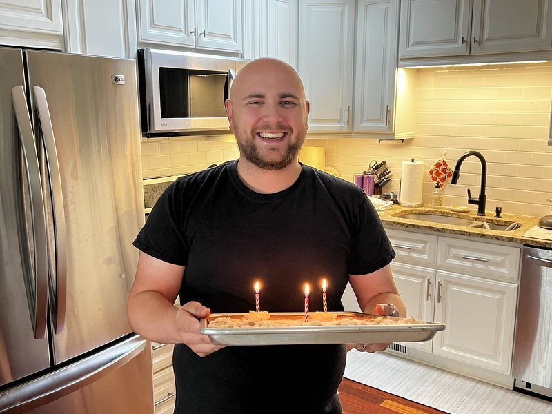 Alex in his kitchen holding up a pan of creatively frosted birthday banana bars with lit candles
