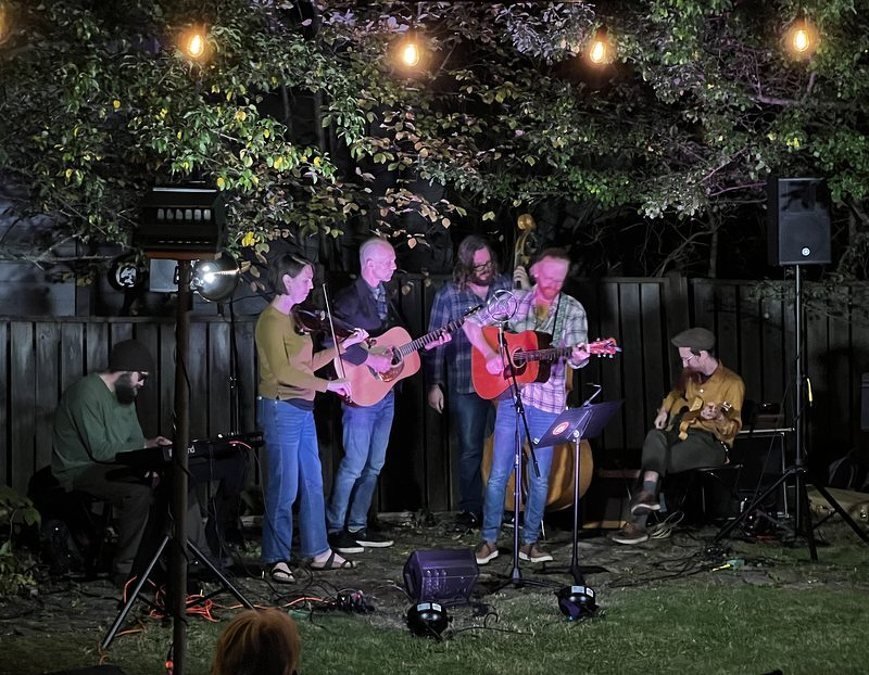 A full band of fiddle, guitars, bass, and keys playing on the grass at dusk under bistro lights and a canopy of trees